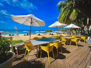 Un patio frente a la playa con sillas amarillas y sombrillas blancas, con vista a una playa arenosa y al océano.