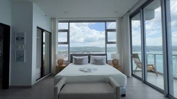 A spacious bedroom with a large bed, a balcony with a view of the sea, and a sliding door leading to another room.