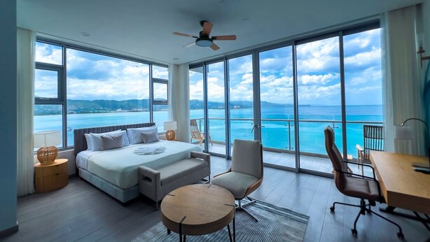 Quarto de hotel moderno com uma cama grande, uma mesa, uma cadeira e vista para o mar.