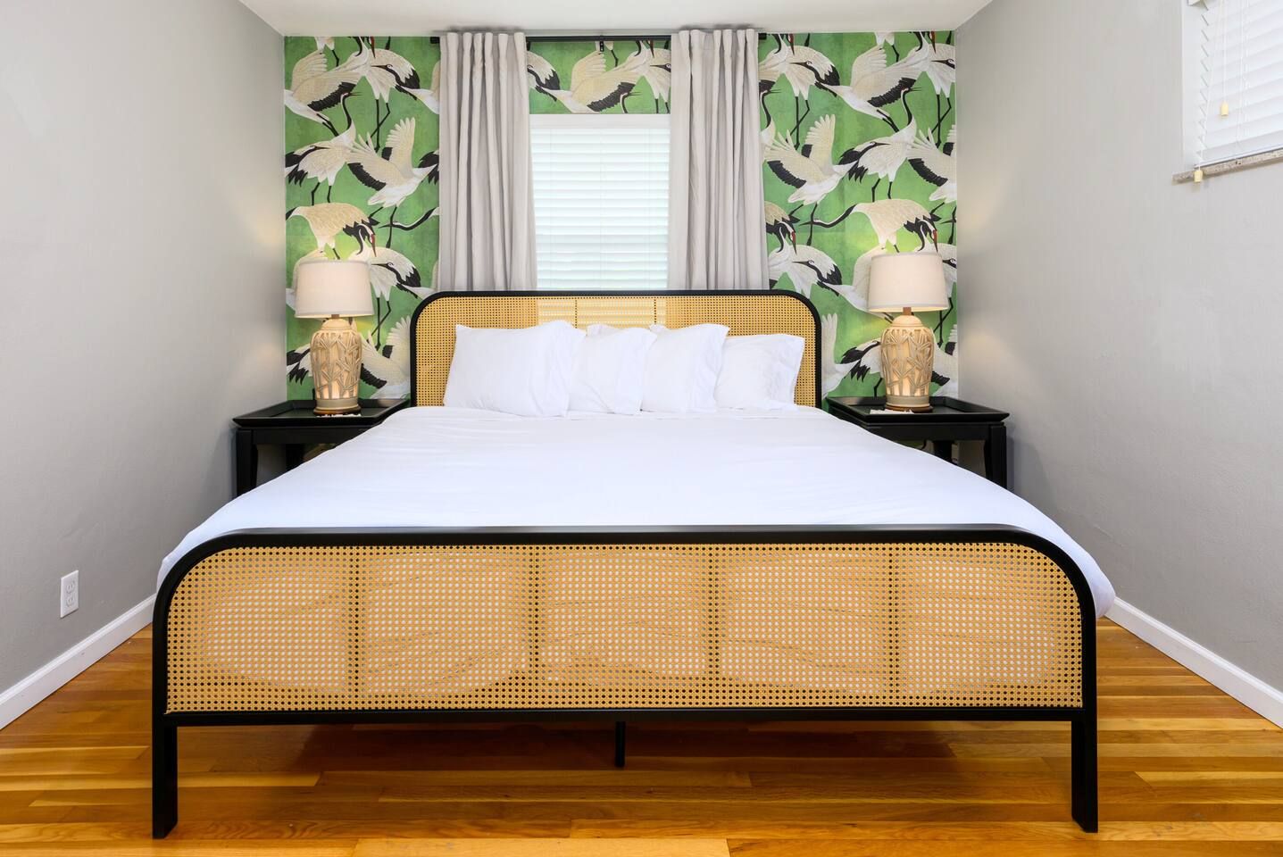 Una cama con cabecera trenzada, dos mesitas de noche con lámparas y una pared con estampado de pájaros verdes y blancos.