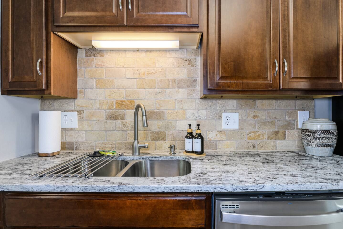 A kitchen with a granite countertop, stainless steel sink, wooden cabinets, and a stone backsplash.