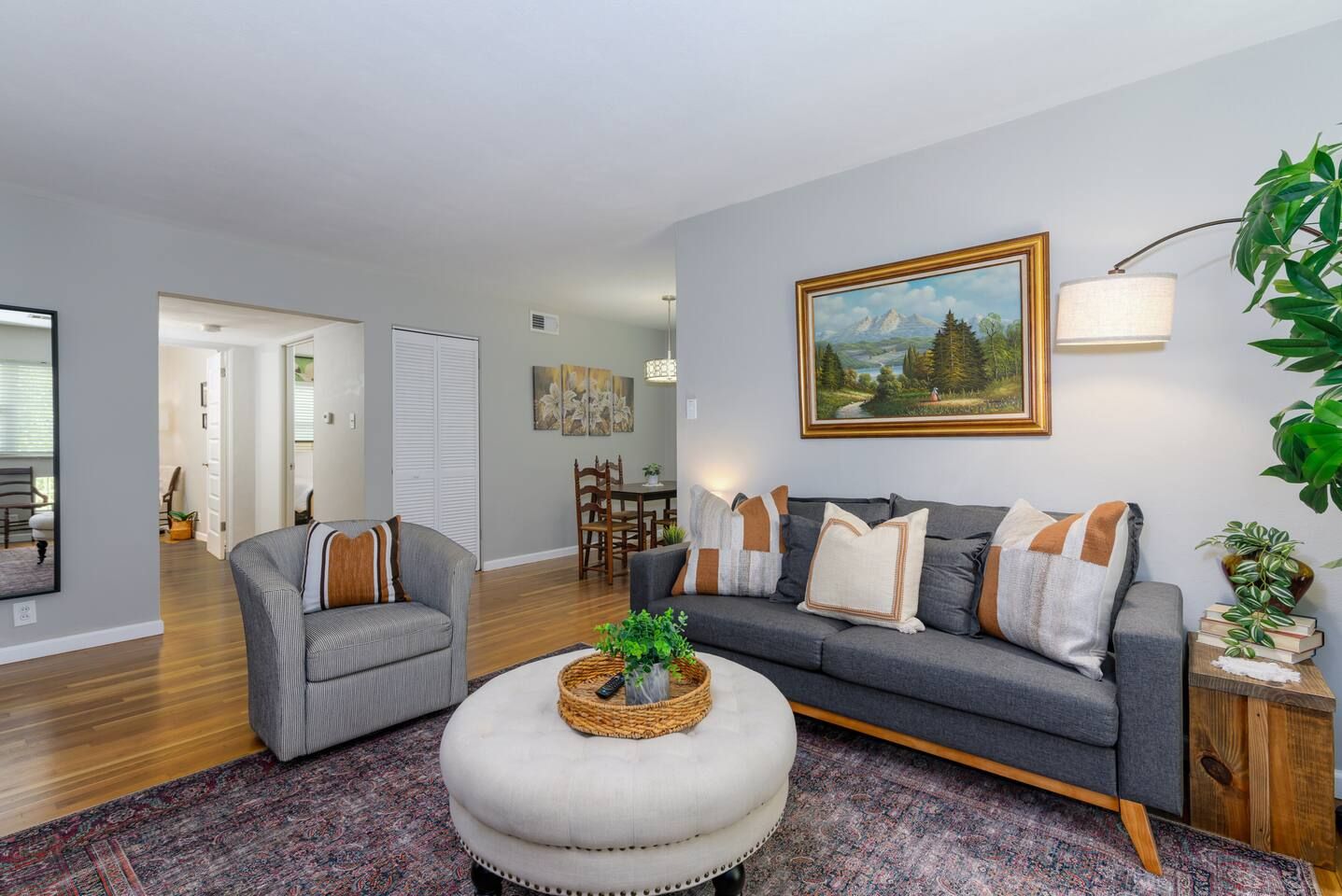 Una sala de estar moderna con un sofá gris, un pouf redondo y un comedor al fondo.