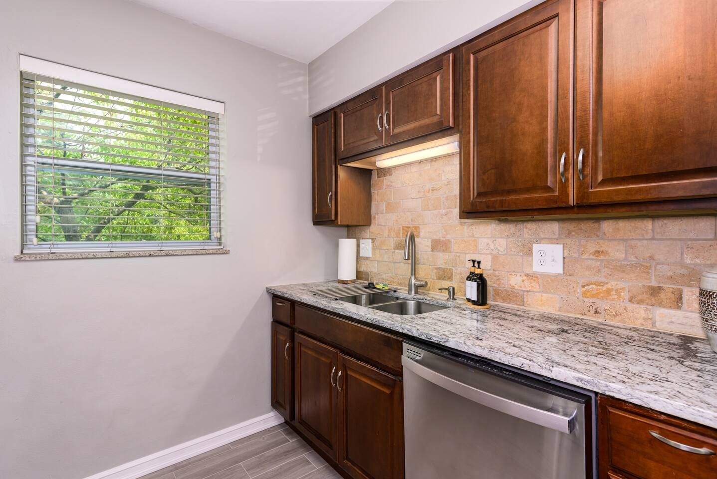 A kitchen with wooden cabinets, a granite countertop, a stainless steel dishwasher, and a window with blinds.