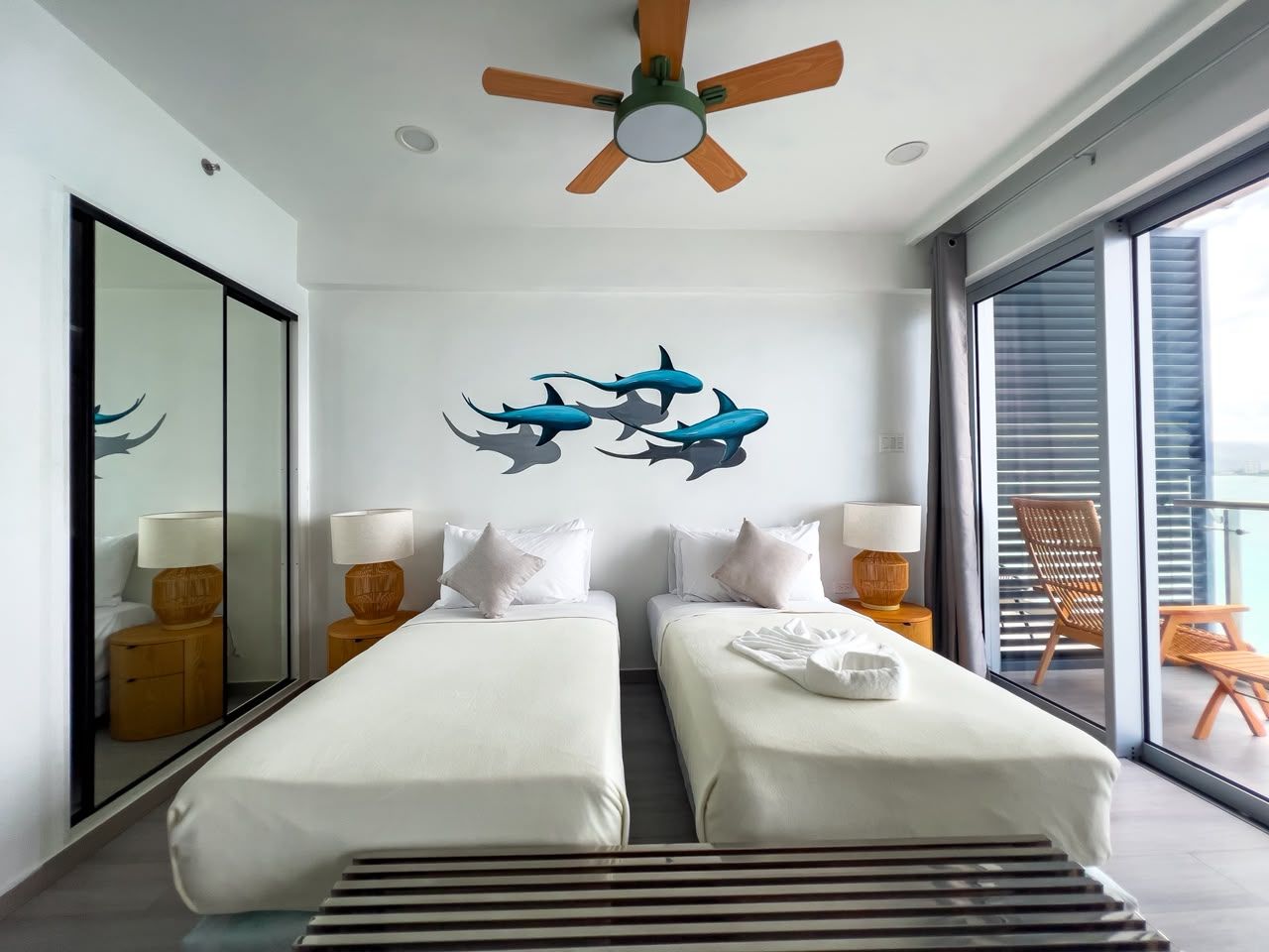 A hotel room with two beds, a ceiling fan, a large window, and a balcony with outdoor furniture.