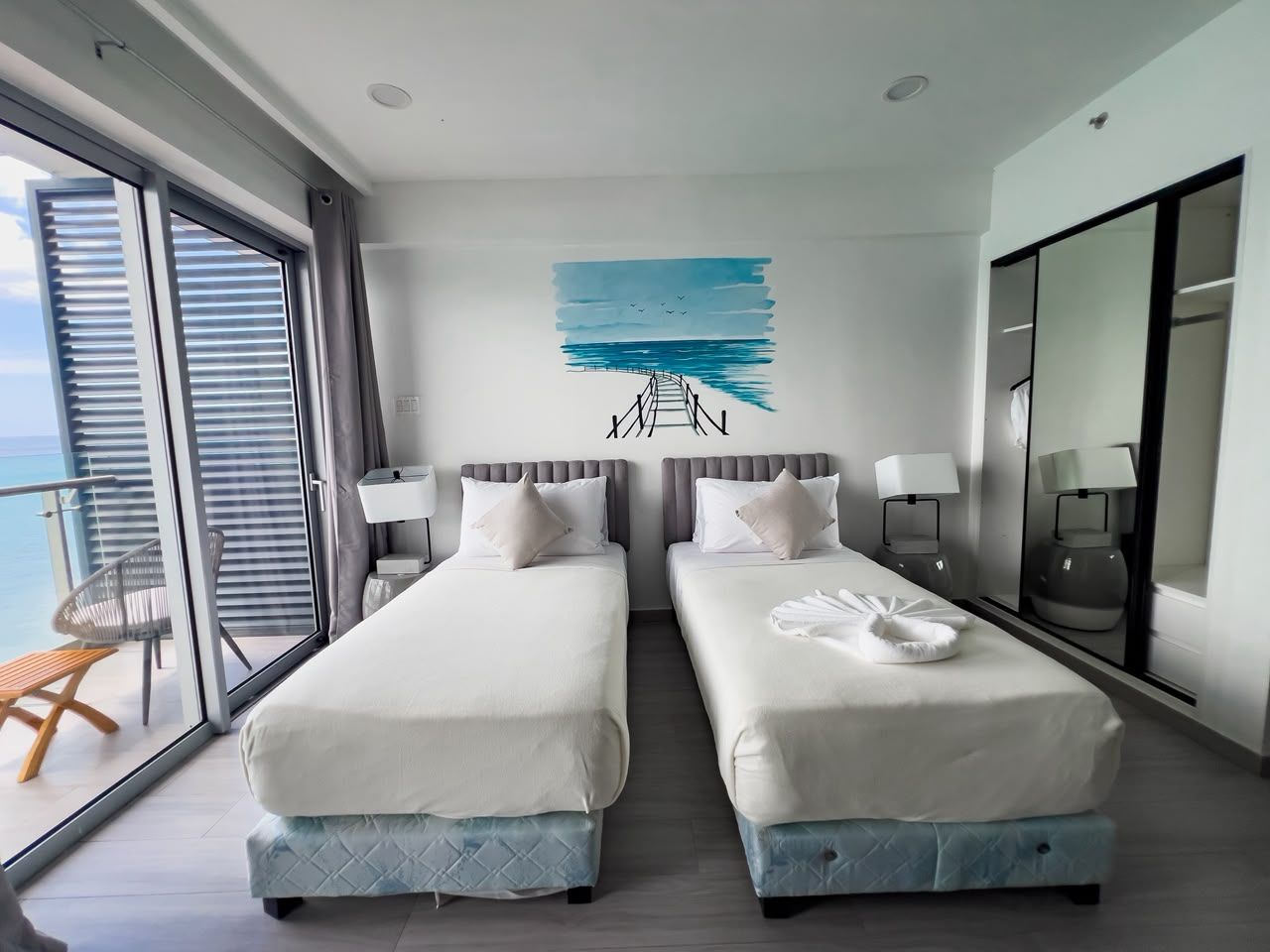 A hotel room with two beds, a large window, a balcony, and a wall art depicting a seascape.
