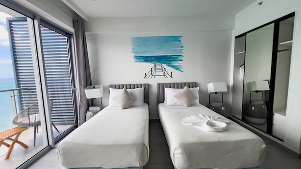 Habitació d'hotel amb dos llits, una finestra gran, un balcó i una obra d'art mural que representa un paisatge marítim.
