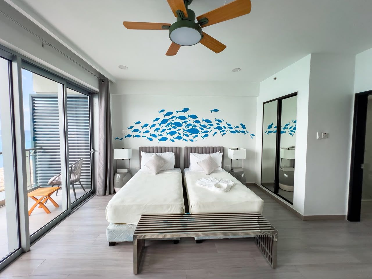 A hotel room with two beds, a ceiling fan, a large window with blinds, a balcony with a table and chairs, and a wall mural of fish.