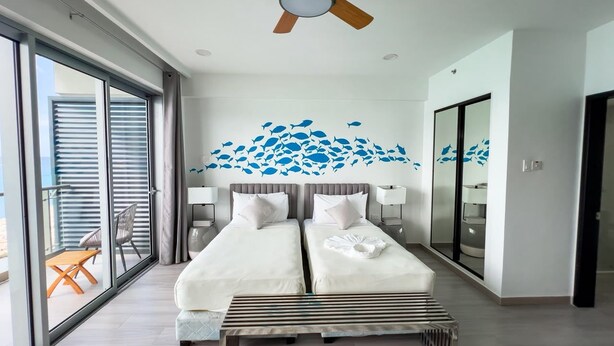 Habitació d'hotel amb dos llits, un ventilador de sostre, una finestra gran amb persianes, un balcó amb taula i cadires, i un mural al mur amb motius de peixos.