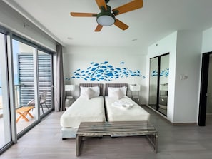 A hotel room with two beds, a ceiling fan, a large window with blinds, a balcony with a table and chairs, and a wall mural of fish.