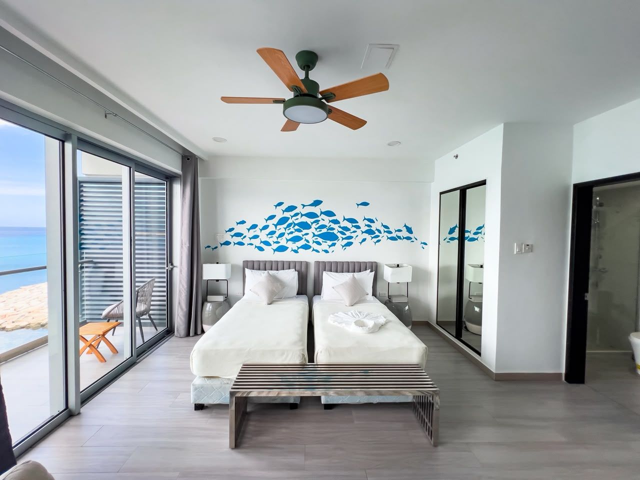 A modern bedroom with a large bed, a ceiling fan, and a view of the beach through floor-to-ceiling windows.