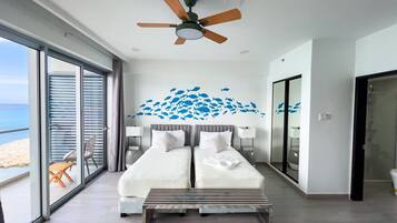A modern bedroom with a large bed, a ceiling fan, and a view of the beach through floor-to-ceiling windows.