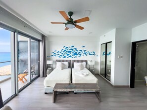 Ein modernes Schlafzimmer mit einem großen Bett, einem Deckenventilator und Blick auf den Strand durch raumhohe Fenster.