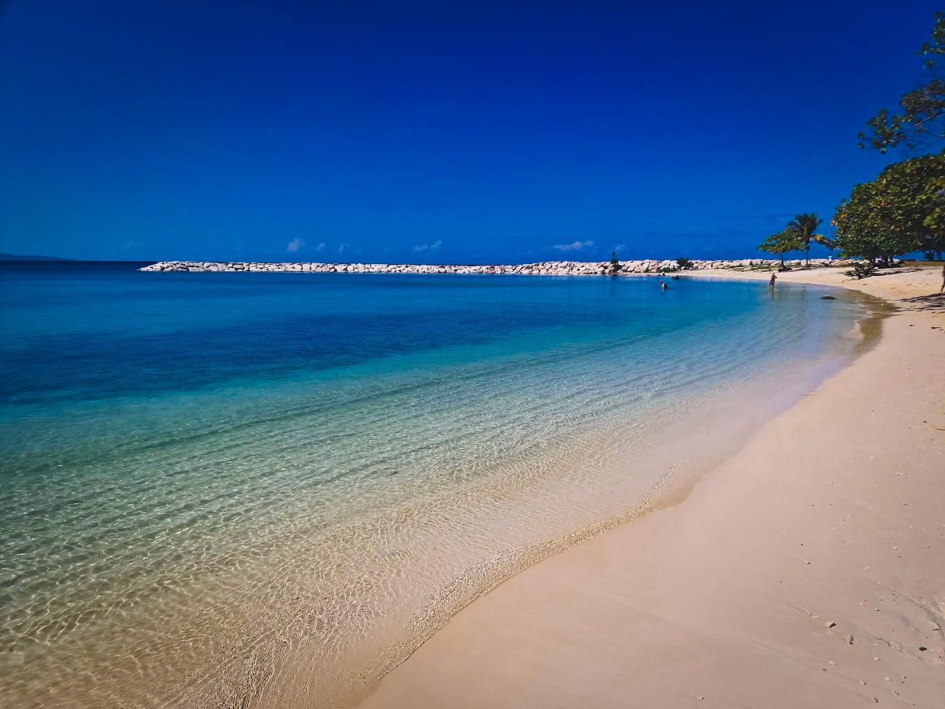 A beach with clear water, a sandy shore, and a rocky outcrop in the distance.