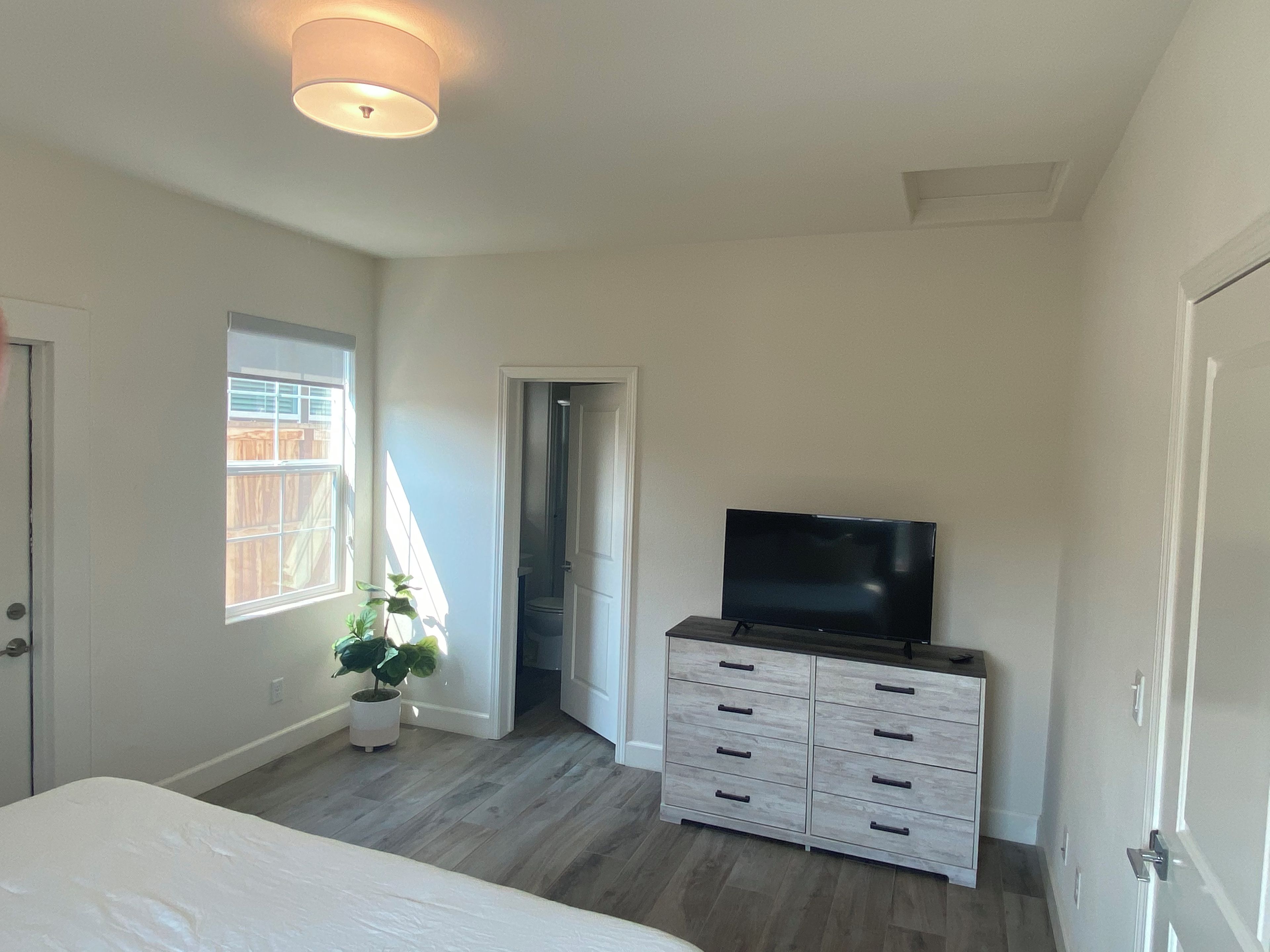 A bedroom with a bed, a dresser, a television, a window, and a door leading to a bathroom.