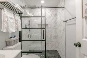 Un baño moderno con cabina de ducha de vidrio, baldosas de mármol blanco, un lavabo blanco y una cesta de toallas.