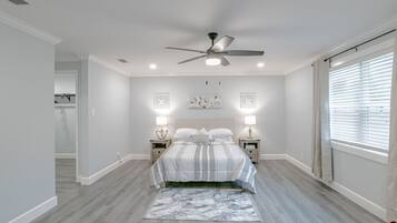 Un dormitorio con una cama, dos mesitas de noche, un ventilador de techo y un amplio ventanal con persianas.