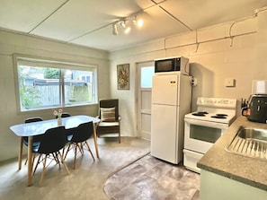 A compact kitchen with a refrigerator, stove, and microwave. There is a dining table with chairs and a window providing natural light. - Big Beachside Bach/1 min to Sand & Sea/Sleeps 10 (Tauranga)