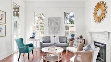 Una sala de estar con un sofá gris, un sillón verde azulado, sillas de mimbre, una mesa de centro redonda y una chimenea blanca.