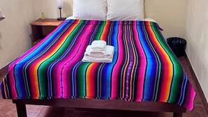 Uma cama com colcha listrada e colorida, almofadas brancas e uma pequena mesa de cabeceira com abajur e uma garrafa.