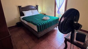 Quarto com cama, mesa de cabeceira, ventoinha na parede e janela com cortinas.