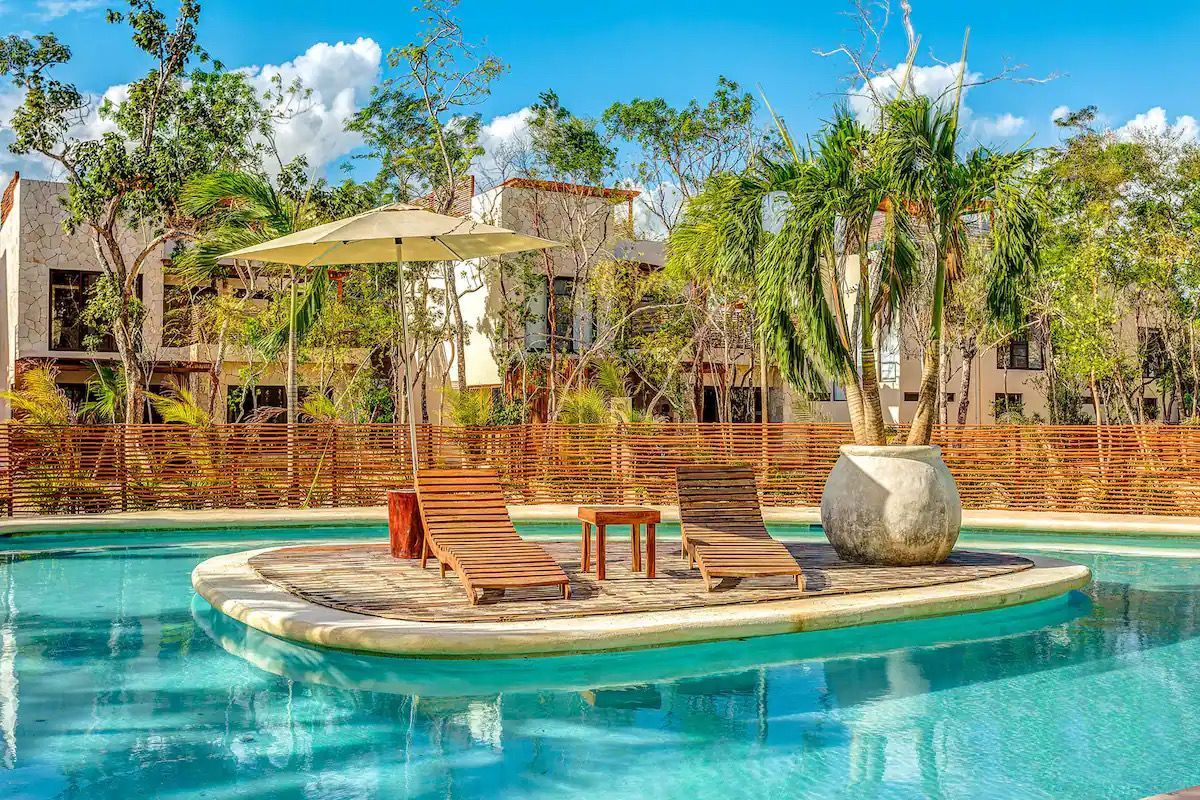 A pool with lounge chairs and a table, surrounded by a wooden fence and tropical vegetation.