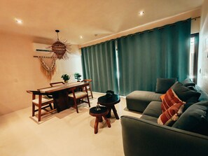 A living room with a sofa, a dining table with chairs, a hanging light fixture, and a view of the outdoors through a window with curtains. - Peaceful Luxury Ground-Floor Retreat, 2BR, 2BA, Selva Norte Tulum (Tulum)