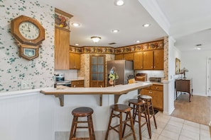 A kitchen with wooden cabinets, a central island with stools, a refrigerator, and a wall clock.