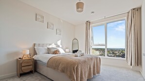 Ein Schlafzimmer mit großem Fenster, einem Bett mit flauschiger Decke, einem Nachttisch mit Lampe und Blick auf eine Stadtlandschaft.