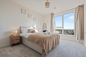 Et soveværelse med et stort vindue, en seng med et blødt tæppe, et natbord med en lampe, og udsigt over bybilledet.