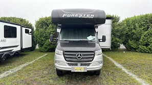 A white camper van with a black camper shell labeled 'FORESTER BY FOREST RIVER' parked on grass with trees in the background. - Luxury Mercedes 25 Foot RV w/ Full Bathroom + WiF/TV close to the Poconos (Blairstown)
