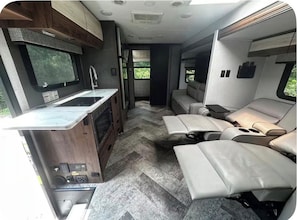 A compact RV interior with a kitchenette, a seating area with cushioned seats, and a window providing a view of greenery. - Luxury Mercedes 25 Foot RV w/ Full Bathroom + WiF/TV close to the Poconos (Blairstown)