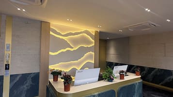 Un mostrador de recepción con una computadora portátil, plantas en macetas y una pared con diseño ondulado.