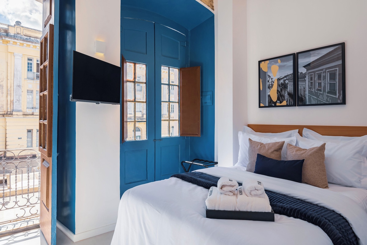 Habitación de hotel con una cama grande, un televisor de pantalla plana montado en la pared y un balcón con vista a la ciudad.