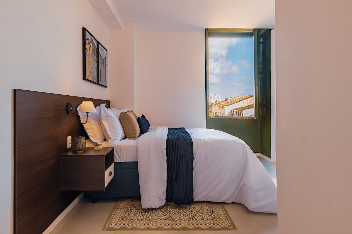 Un dormitorio con cama, mesita de noche, lámpara y vistas a un edificio a través de una ventana.