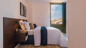 Un dormitorio con cama, mesita de noche, lámpara y vistas a un edificio a través de una ventana.