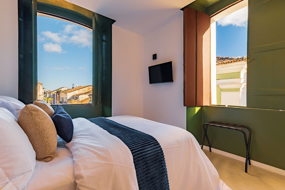Habitación de hotel con cama, televisor montado en la pared y ventana con vistas a edificios.