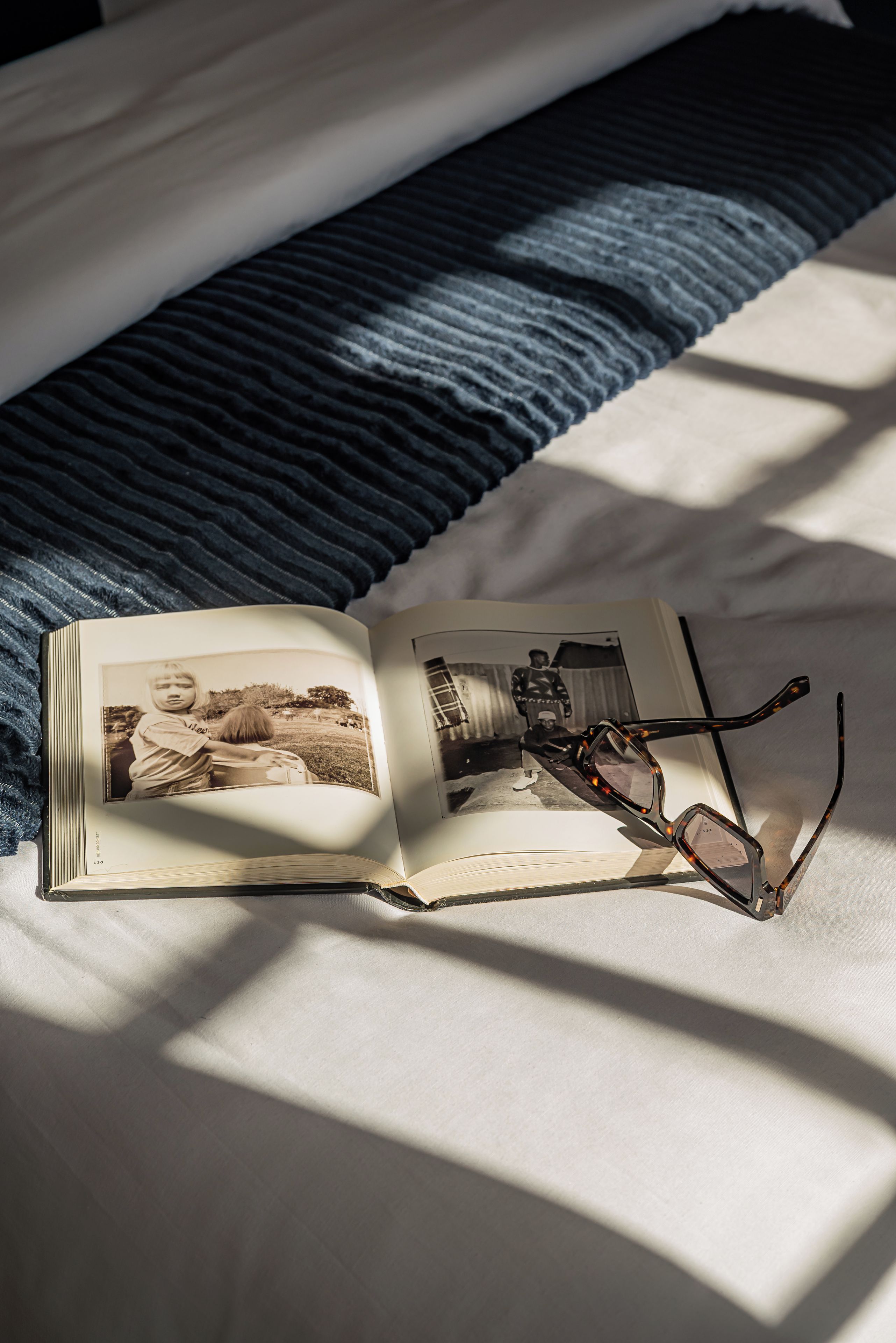 Een open boek met een foto op één bladzijde en een paar brillen op de andere, neergelegd op een bed met een blauwe deken.