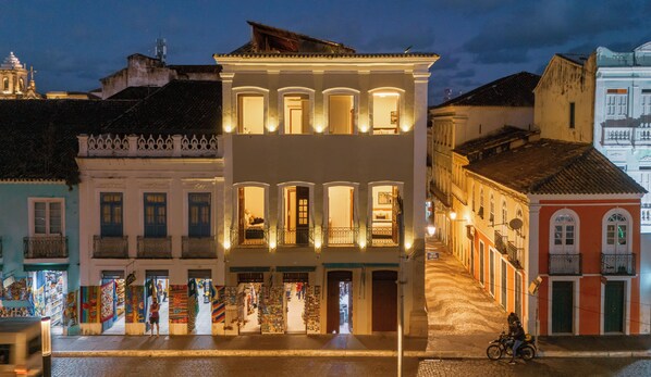 A multi-story building with illuminated windows and balconies, a street in front with shops, and a motorcycle parked on the sidewalk.