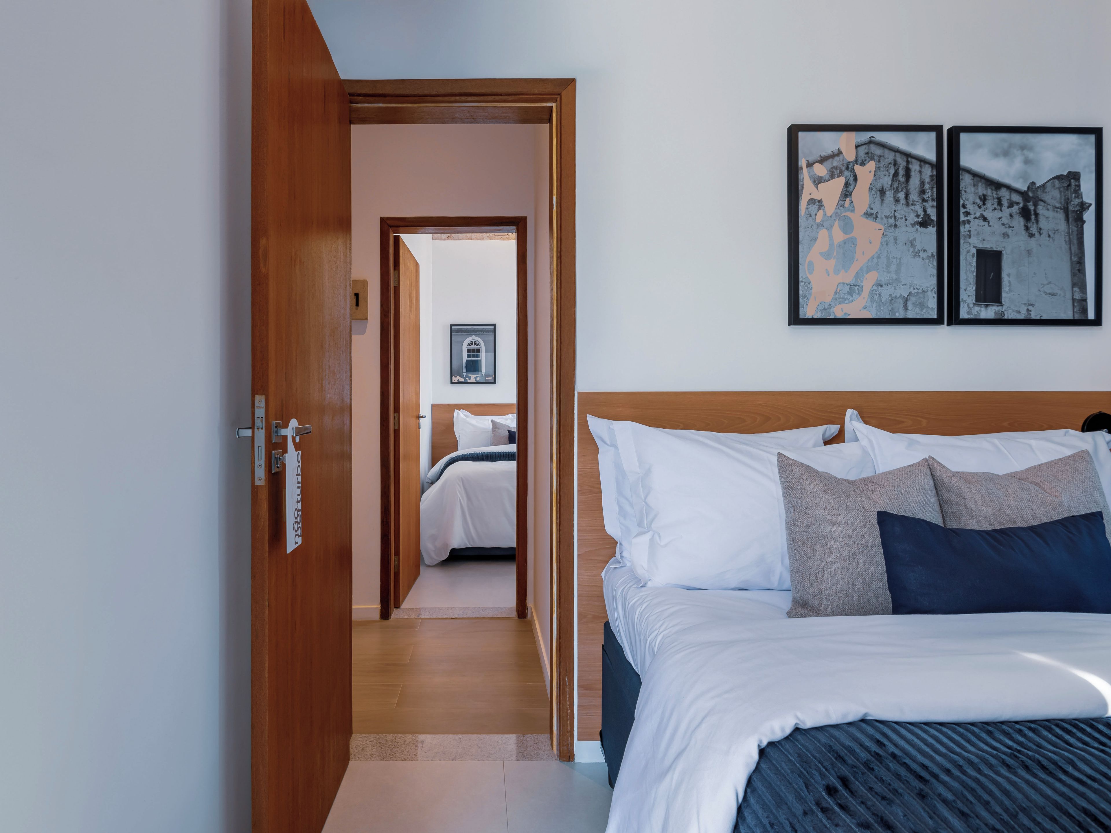Una habitación de hotel con una puerta de madera, una cama con ropa de cama blanca y azul oscuro, y dos cuadros en la pared.