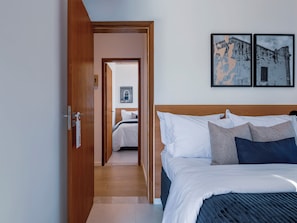 Una habitación de hotel con una puerta de madera, una cama con ropa de cama blanca y azul oscuro, y dos cuadros en la pared.