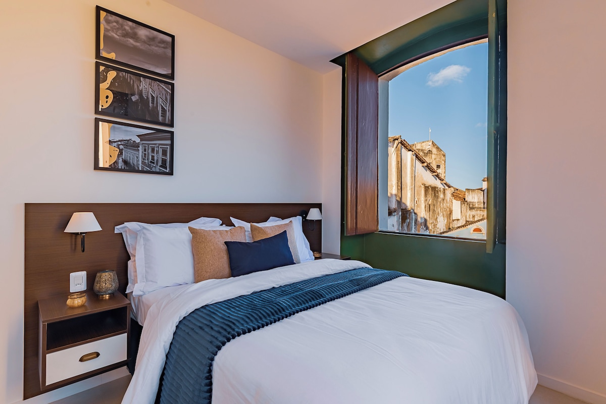 Ein Hotelzimmer mit einem großen Bett, einem Nachttisch mit Lampe, einem Wandregal und einem Fenster mit Blick auf Gebäude.