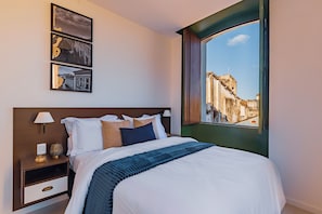 Una habitación de hotel con una cama grande, una mesita de noche con lámpara, un estante en la pared y una ventana con vistas a edificios.