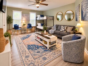 A modern living room with a sofa, armchair, and a coffee table. There is a bunk bed in the background, a large rug, and a ceiling fan. - Dunes & Beach Getaway - Sleeps 10 - Room for the Whole Crew (Portage)