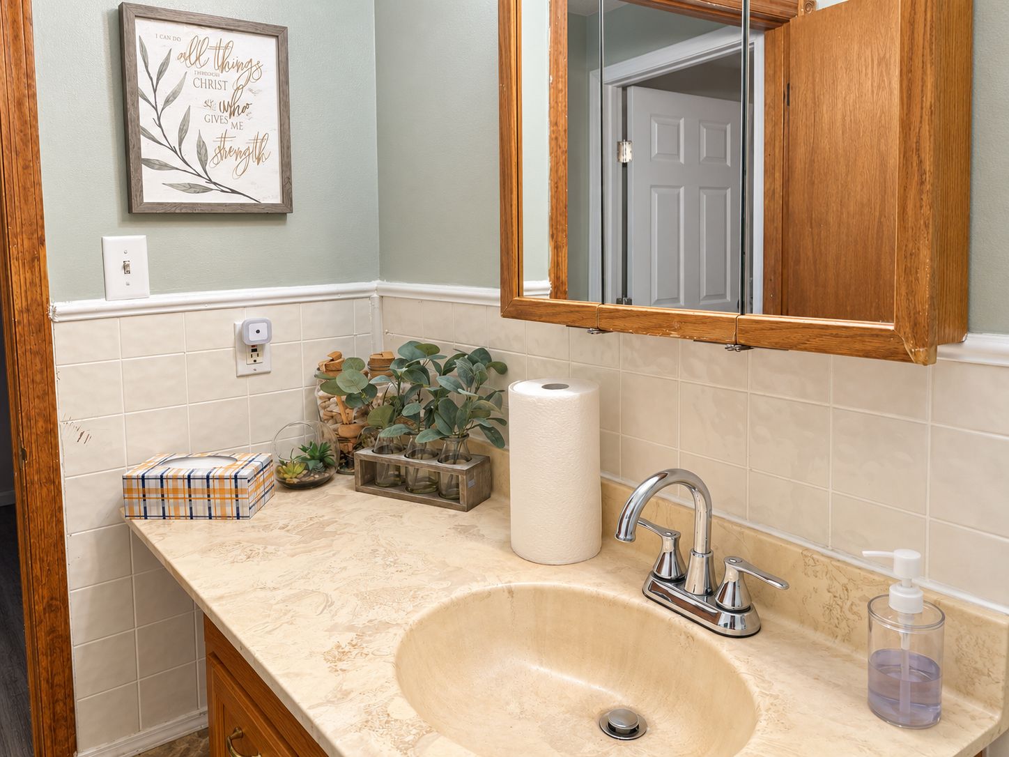A bathroom with a beige sink, a mirror with a wooden frame, a toilet paper roll, and a small plant on the counter.
