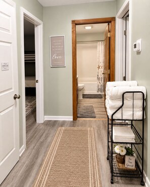 A hallway with a wooden door, a framed inspirational quote, a metal towel rack with a sign, and a patterned rug. - Dunes & Beach Getaway - Sleeps 10 - Room for the Whole Crew (Portage)