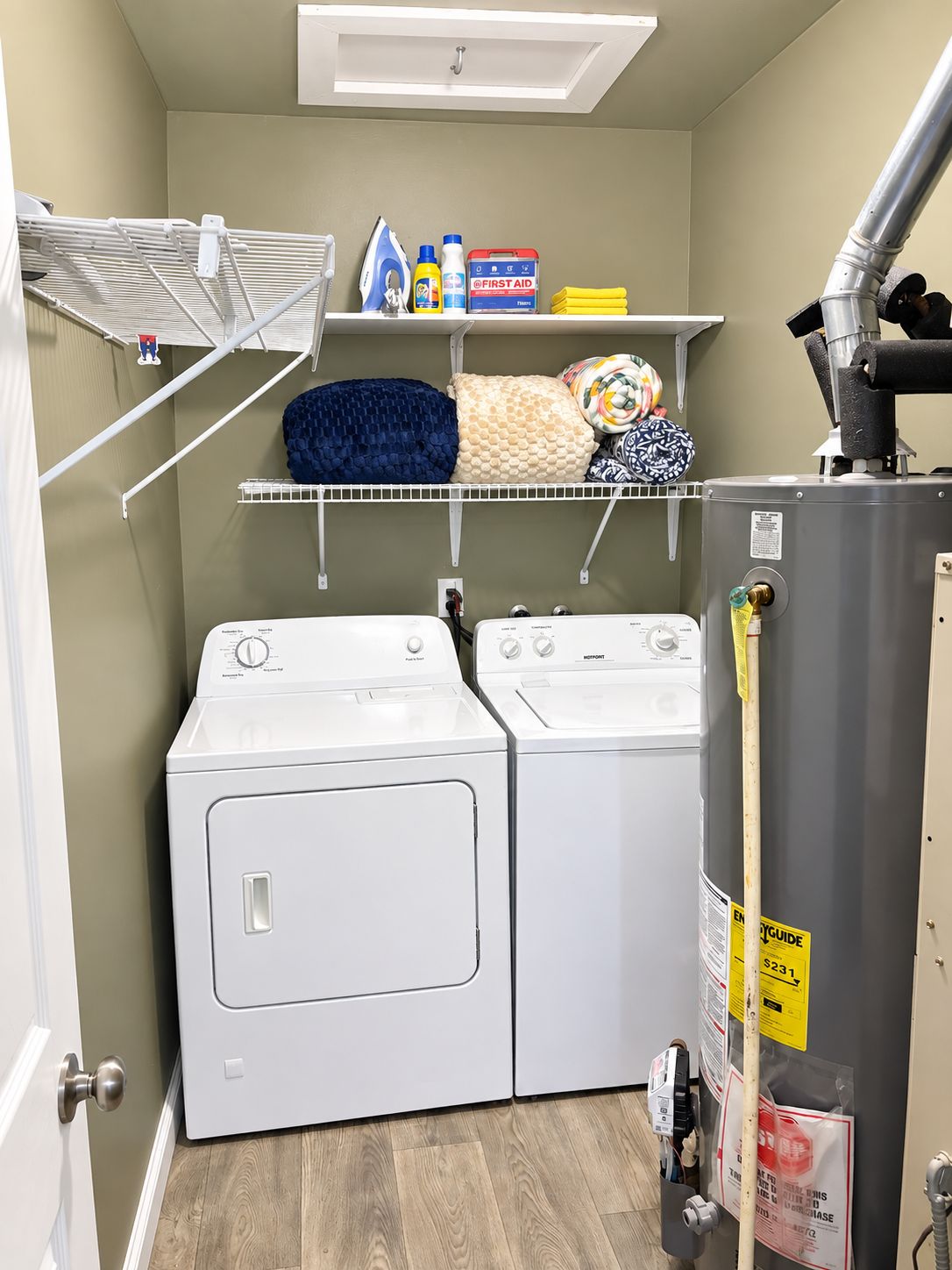 A laundry room with two front-loading washing machines, a shelf with laundry supplies, and a vent pipe.