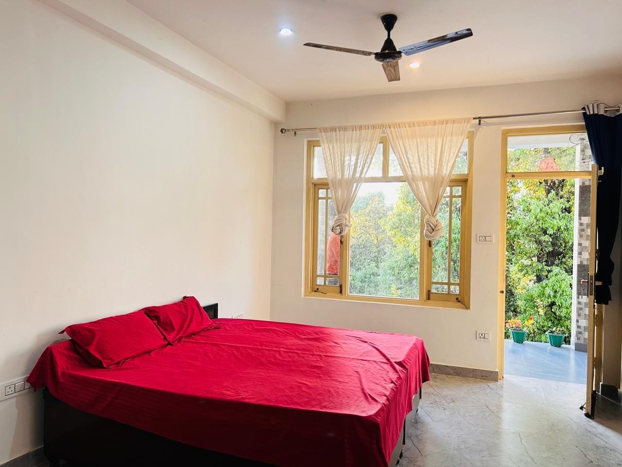 A bedroom with a bed, a ceiling fan, a window with sheer curtains, and a view of greenery outside.