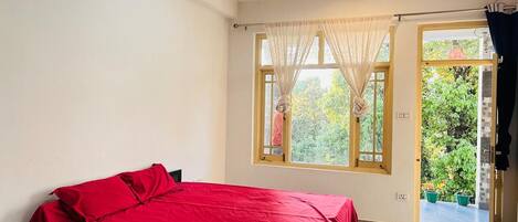 A bedroom with a bed, a ceiling fan, a window with sheer curtains, and a view of greenery outside.