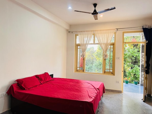 A bedroom with a bed, a ceiling fan, a window with sheer curtains, and a view of greenery outside.