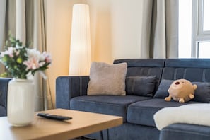 En moderne stue med en mørkeblå sofa, en beige pude, en blød bamse, en vase med blomster og en fjernbetjening på et træbord.